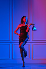 African american model in little dress, fishnet tights holds hand bag under neon lights