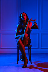 African american woman in dress, fishnet tights sits on chair with hand bag under neon lights