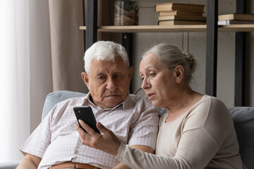 Senior couple discuss mobile app seated on sofa at home, staring at smart phone screen learn new software. Loving wife explains modern device usage to mature husband. Help, tech and older gen concept
