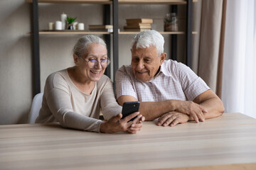 Smiling older couple sit at table staring at smart phone screen, enjoy new mobile application, lead...