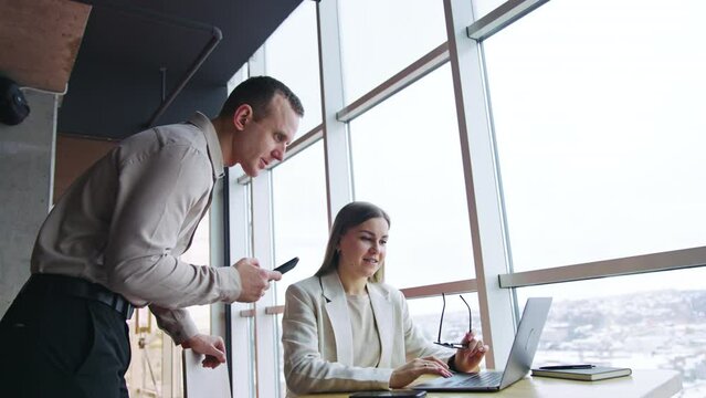 Office colleagues collaborating at work. Teammates look at laptop and discuss business issues. Low angle view. Panoramic windows at backdrop.