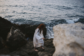 A woman in a white dress stands barefoot on the rocks by the ocean