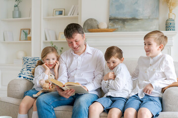 Caring father reads book to his little daughter and his sons hugs, sits on cozy sofa at home, family spends their free time together, loving dad with children is engaged in educational activities