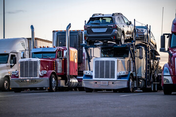 Big rigs semi trucks tractors with car hauler and refrigerator and dry van semi trailers standing in row on the truck stop industrial parking lot for the rest at twilight time © vit