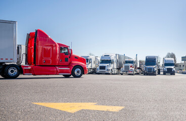 Bright red big rig semi truck with extended cab transporting cargo in dry van semi trailer driving on the truck stop parking lot with direction of movement arrow and row of another semi trucks © vit