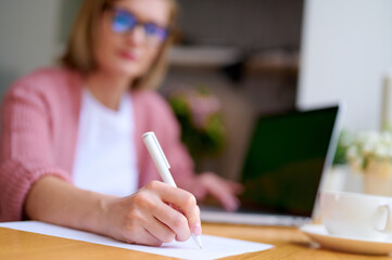 Selective focus woman blogger working at laptop at home holds a pen in her hands and make a plan...