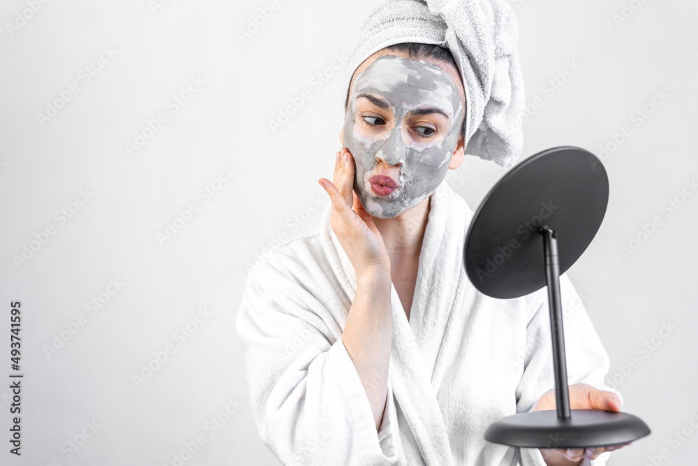 Sticker Young woman with a clay mask on her face in front of a mirror.