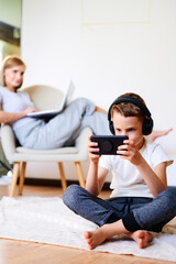 Child plays mobile game on the phone while the mother is working on the laptop. Schoolboy holds mobile phone in his hands. Distance learning. Cybersport.
