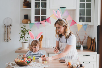 charming woman and her two-year-old son in bunny ears paint Easter eggs with paints sitting at the table on the kitchen