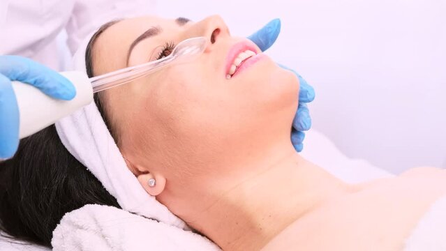 Young beautiful smiling woman receiving facial electric apparatus darsonval procedure in a beauty clinic by a professional cosmetologist.