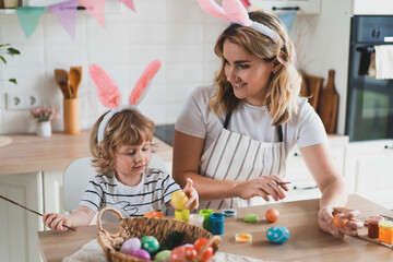 Fototapeta premium charming woman and her two-year-old son in bunny ears paint Easter eggs with paints sitting at the table on the kitchen
