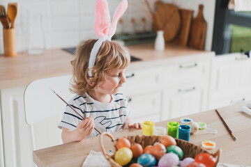 Cute two-year-old boy in bunny ears paint Easter eggs with paints sitting at the table on the kitchen