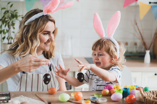 charming woman and her two-year-old son in bunny ears paint Easter eggs with paints sitting at the table on the kitchen