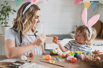 charming woman and her two-year-old son in bunny ears paint Easter eggs with paints sitting at the table on the kitchen