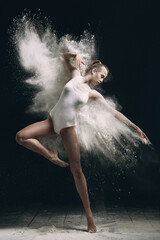 Young beautiful girl in a bodysuit in an explosion of white powder isolated on a dark black background. The concept of energy, power, movement. concept for advertising. Splashes of white paints on a d