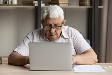 Serious older man sit at table use laptop, looking focused, read information, typing letter, lead correspondence to support service. Elderly people and modern tech, internet, e-services usage concept