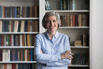 Happy middle-aged woman standing with arms crossed on bookshelves background, staring at camera...