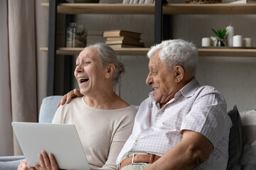 Happy mature couple spend time use laptop relax on sofa in living room, put computer on laps watch movie laughing enjoy comedy at home. Older generation and modern tech usage, leisure and fun concept