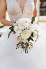 The bride in a white wedding dress holds a bouquet of white peonies and greenery in her hands