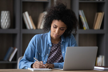 Serious young African Black student girl writing notes at laptop, studying in library, attending online class, watching learning virtual webinar on computer, listening to teachers tutorials