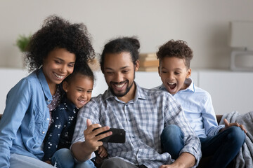 Happy joyful Black family with beloved preschool kids taking selfie on smartphone. Young couple of parents and siblings resting on couch, posing for self portrait on cellphone, having fun, laughing