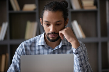 Focused Afro American adult student using laptop in library, preparing for exam, studying online,...