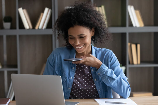 Happy African entrepreneur girl making mobile phone call, talking on speaker, recording audio message for online chat, giving voice command on cellphone to AI virtual assistant