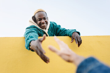 Black African Man helping caucasian friend offering hand to climb wall. Concept of, friendship, solidarity and support - obrazy, fototapety, plakaty