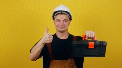 Happy worker in uniform and hard hat holding tool box and showing thumbs up