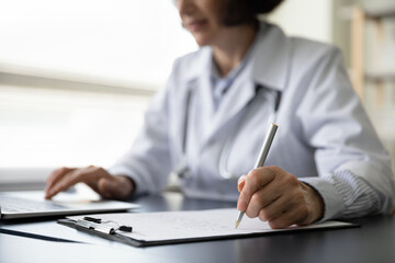 Hands of mature doctor woman filling paper medical records. Practitioner in white coat doing paperwork at workplace with laptop, writing notes, preparing documents, reports, prescription. Close up
