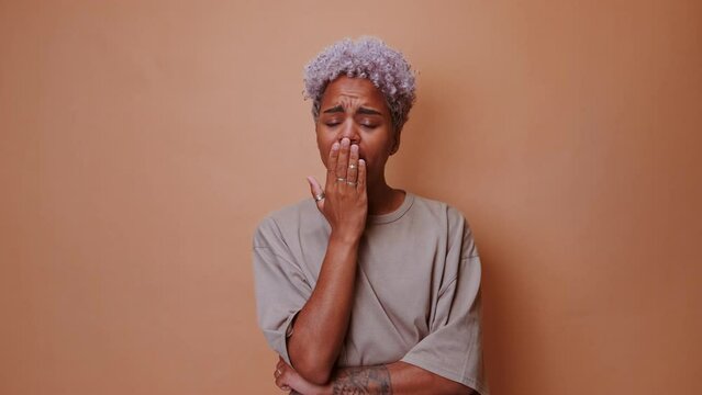 Young tired ethnic African American woman yawns closing eyes and covering mouth with hand wants to sleep after hard day at work she is dressed in simple oversized T-shirt stands on brown background