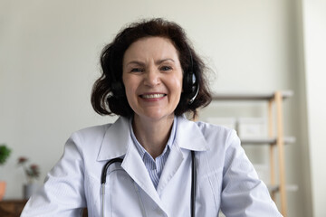Happy mature doctor woman in wireless headphones giving online consultation, medical advice, help on video call, looking at web camera, smiling, laughing. Head shot portrait, screen view
