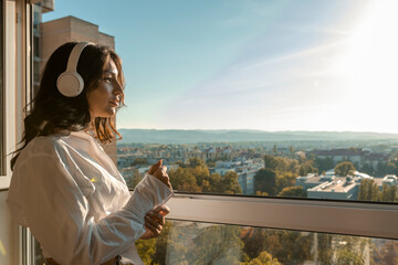 Brunette enjoying sunset on terrace. She is listening to the music via wireless earphones. Casual clothing. Cityscape and mountains in the background.