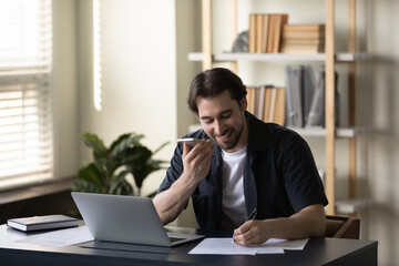 Obraz premium Positive successful business professional man talking on speaker, using mobile phone, recording audio message, giving voice command, writing notes in paper documents on office table