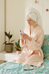 Young happy woman with a towel on her head doing facial skin care procedures. Lies in bed in pajamas in a cozy home with phone. Technology. Social media. Beauty routine. 