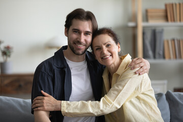 Obraz na płótnie Canvas Happy middle aged mother and grown son hugging with affection, sitting on sofa together, looking at camera, smiling, enjoying family visit, leisure time. Motherhood concept. Head shot portrait