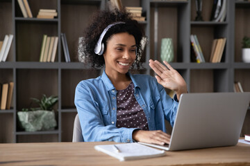 Cheerful Black student girl in wireless headphones waving hand hello at laptop computer, smiling,...