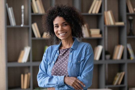 Confident happy young Afro American teacher woman casual posing in college library, looking at camera with toothy smile, keeping hands folded, laughing. African student head shot portrait
