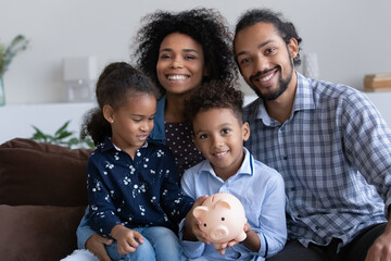 Positive young African parents and little kids showing piggybank with cash, saving money in moneybox, making donation fund, financial reserve for crisis. Happy family bank customers concept