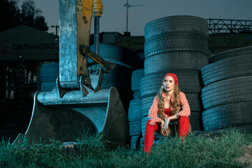 A girl in red sits tired after work near old car tires and an excavator in the evening