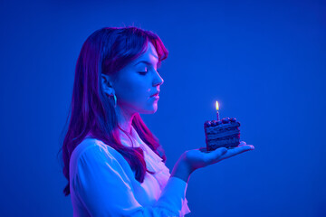 Charming girl blows out candles on a birthday cake. The birthday girl is elegantly dressed. She makes a wish. Happy birthday. ultraviolet background. background trend 2022. neon lights