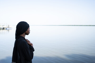 Lonely, troubled young African girl, dressed all in a black shawl and long robe, facing the sea, hands crossed on her chest in an anxious gesture; Loneliness and depression symbol - obrazy, fototapety, plakaty