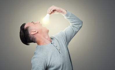A man tries to put a lit electric light bulb in his mouth on grey background.  - obrazy, fototapety, plakaty