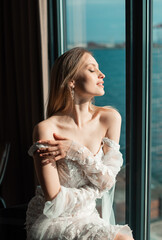 Fashion portrait of bride with a long dress near the window. Waiting for the groom. Wedding interior. Beauty fashion bride at the hotel. Portrait of a beautiful bride. Wedding photography.