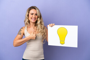 Young Brazilian woman isolated on purple background holding a placard with bulb icon with thumb up