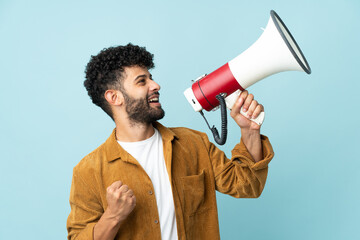 Young Moroccan man isolated on blue background shouting through a megaphone to announce something in lateral position