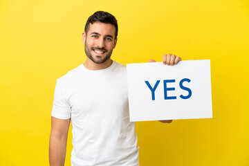 Young handsome caucasian man isolated on yellow background holding a placard with text YES with happy expression