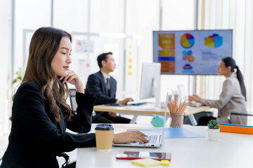 Attractive asian young confident businesswoman sitting at the office table with group of colleagues in the background, working on laptop computer