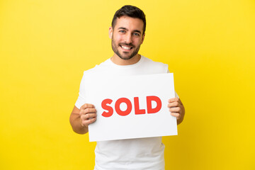 Young handsome caucasian man isolated on yellow background holding a placard with text SOLD with happy expression