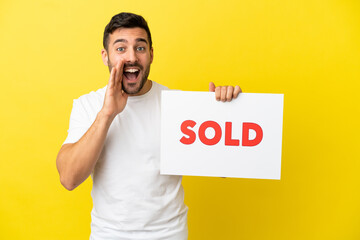 Young handsome caucasian man isolated on yellow background holding a placard with text SOLD and shouting